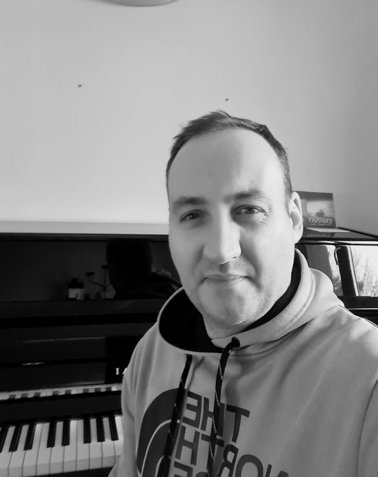 Homme souriant devant un piano, portant un sweat à capuche, en noir et blanc.